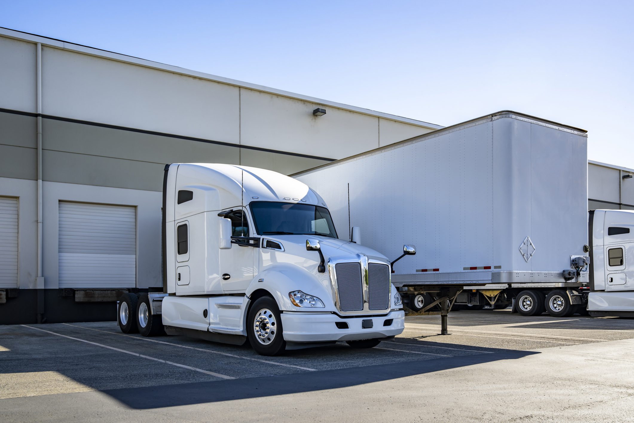 White bonnet industrial big rig semi truck carrier with extended cab for truck driver rest standing side by side with loading cargo dry van semi trailer in warehouse dock gates at sunny day
